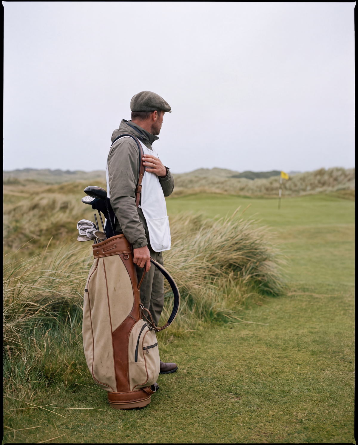 Wes carrying a golf bag at the edge of a links fairway, looking out over the dunes.